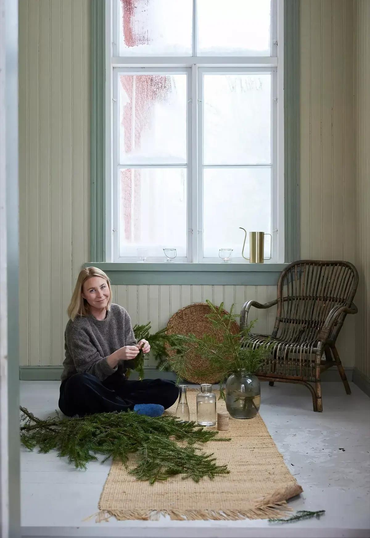 Woman on the floor by the window arranging fir-branch decorations beside a rattan chair