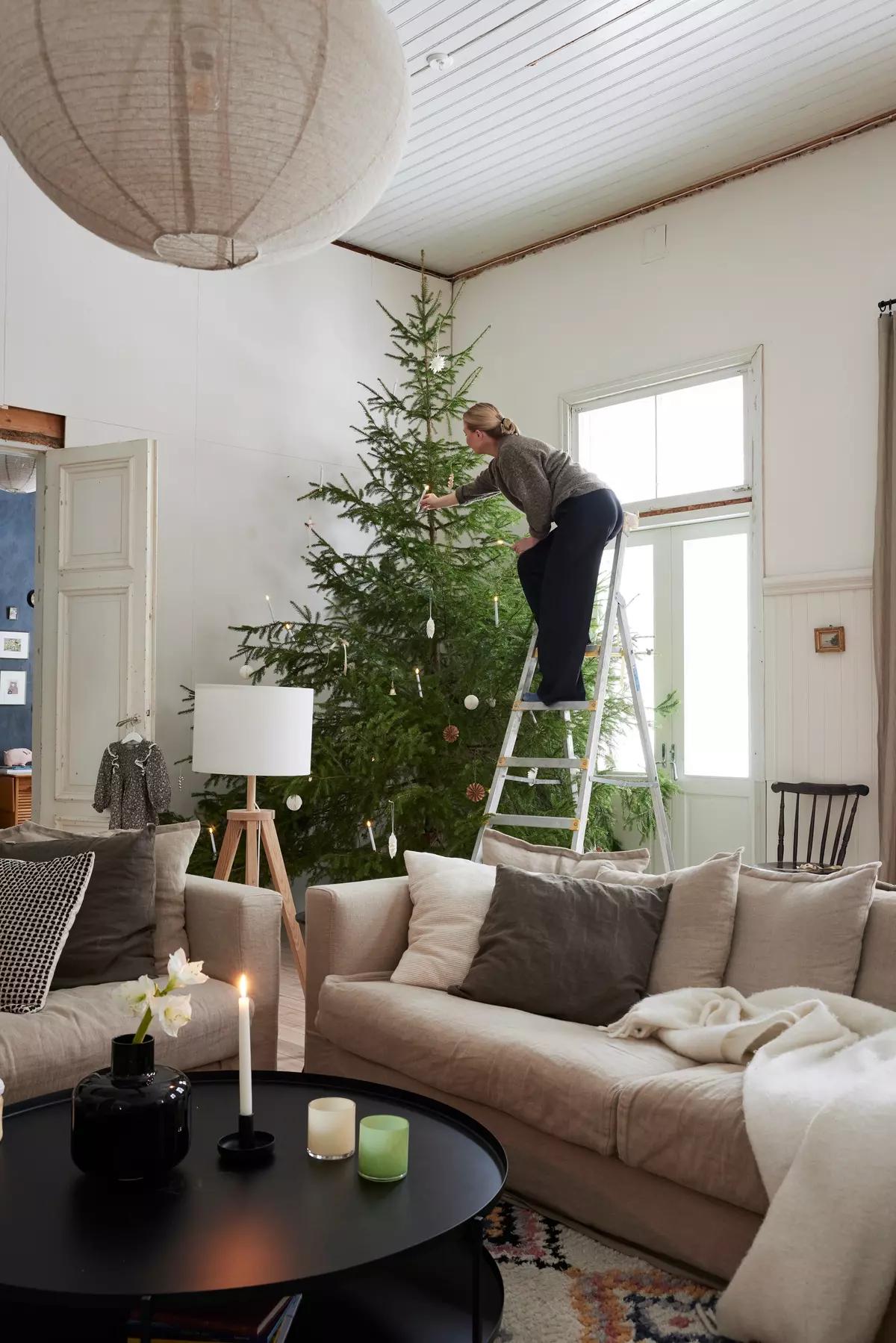 Woman on a ladder decorating the tree in a bright living room with a beige sofa and candles on a round coffee table