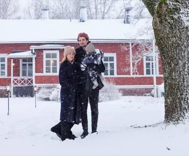 Finnish family holding a child in the snow by a red house with white windows