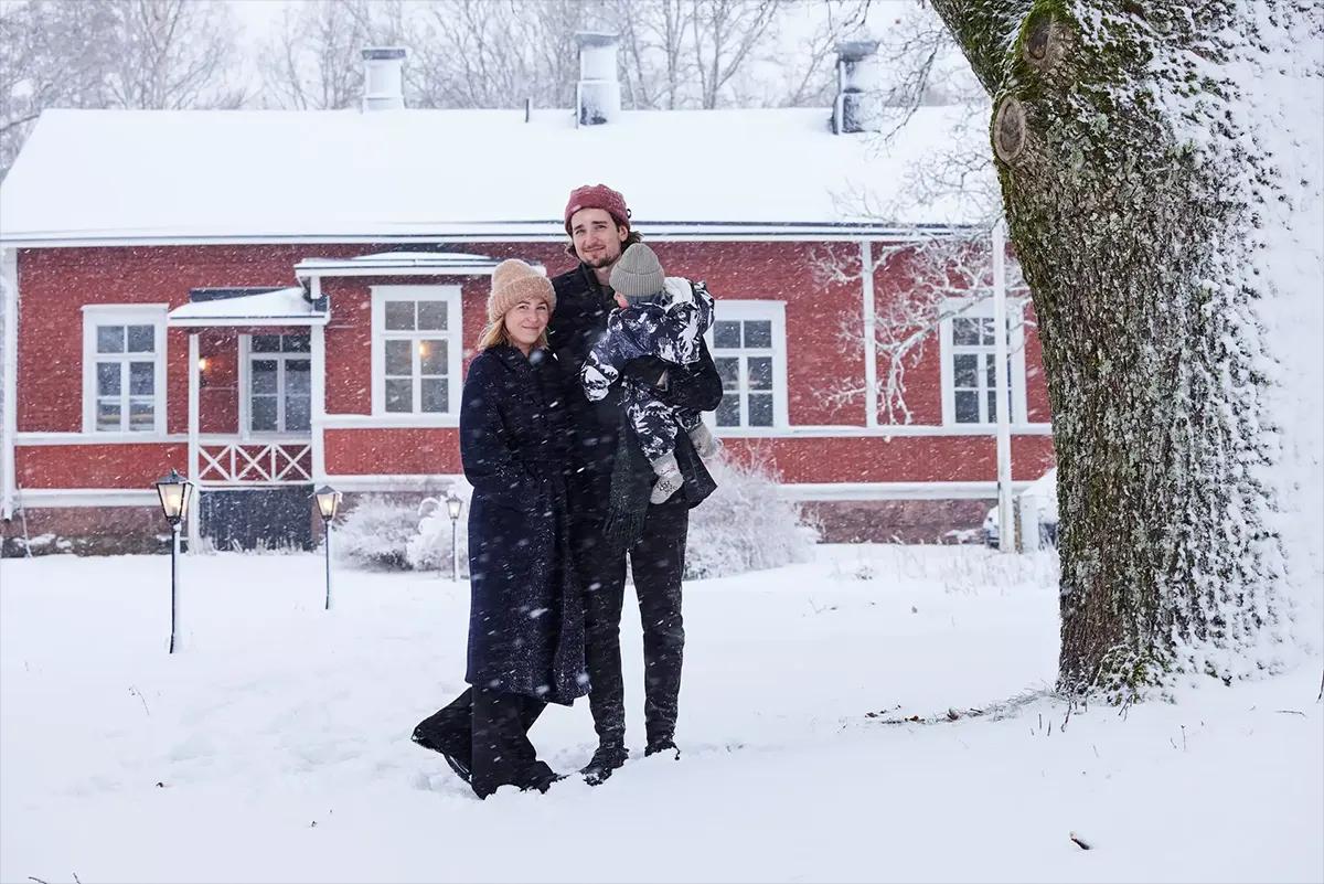 Finnish family holding a child in the snow by a red house with white windows