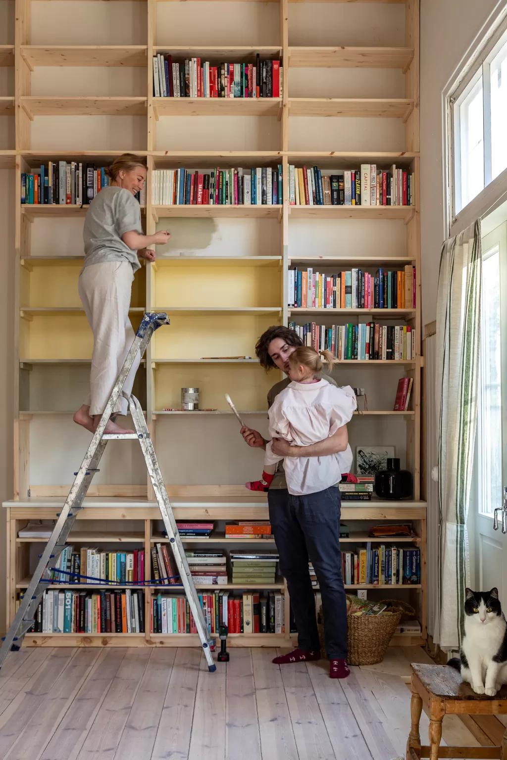 Family painting a bookcase: woman on a ladder, man holding the child, and a cat on a stool