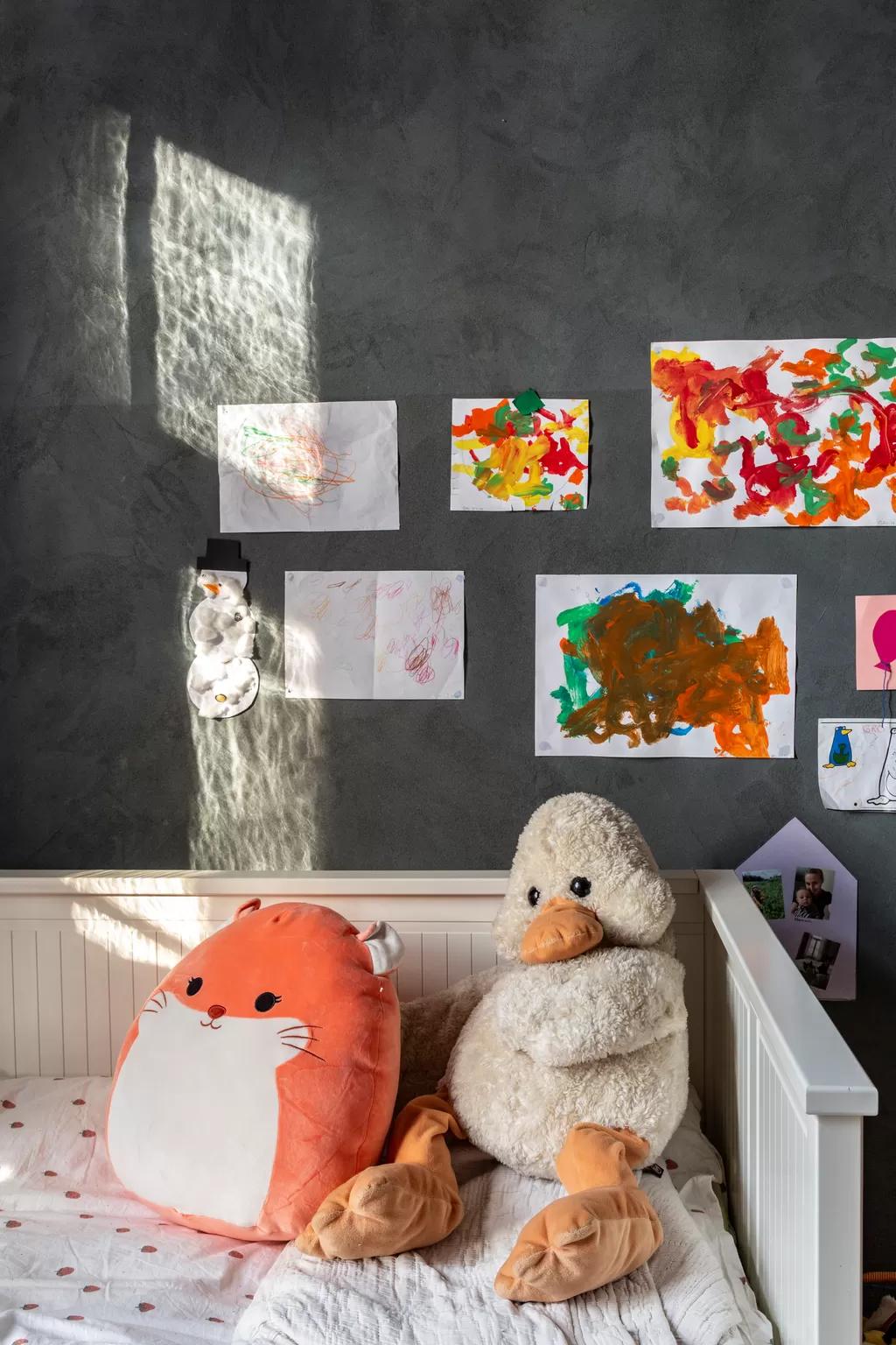 Child’s bed with plush toys and drawings displayed on a grey wall