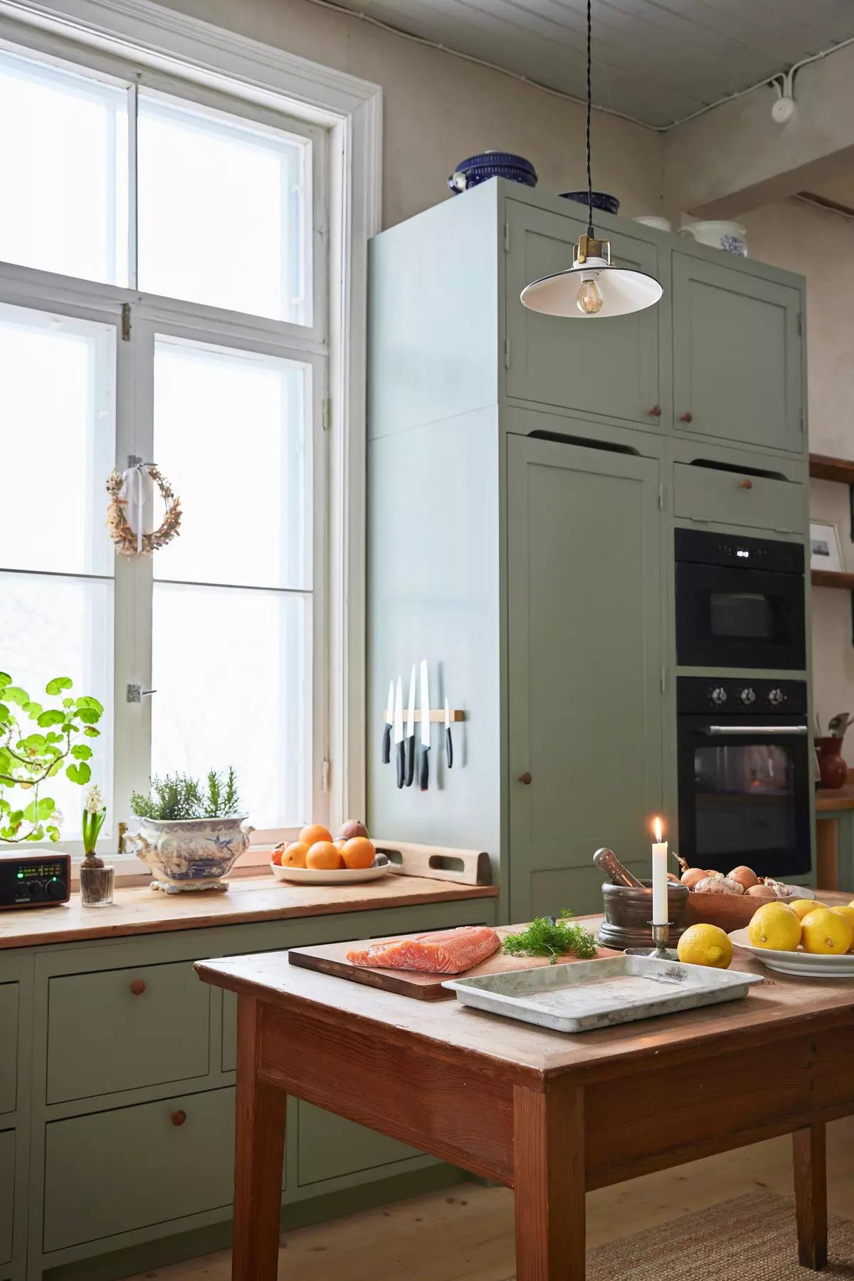 Kitchen with a large window, light-green cabinets and a black oven; salmon fillet, fruit and a candle on the wooden table