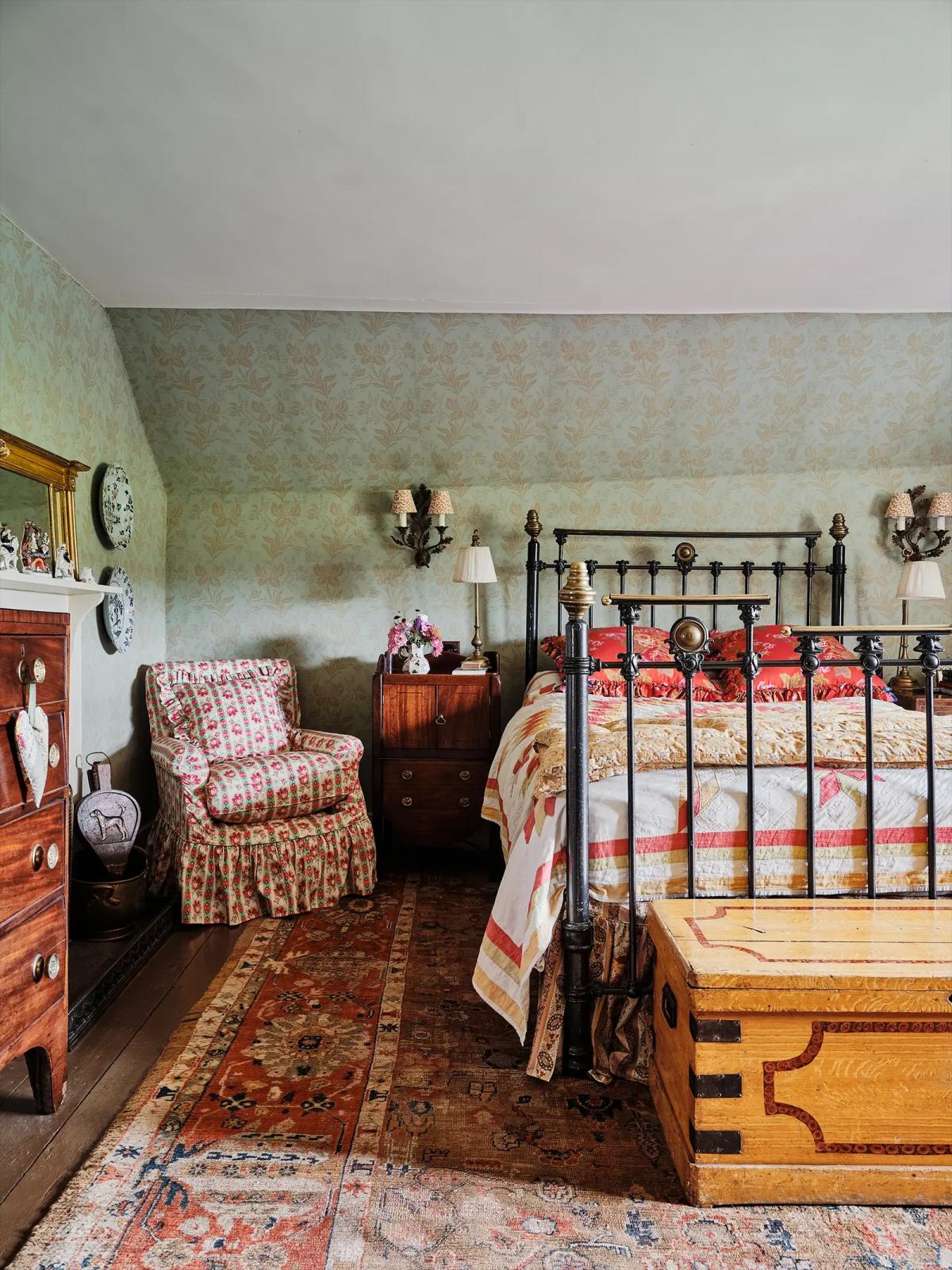 Bedroom with iron bed, armchair, dresser, rugs and floral wallpaper
